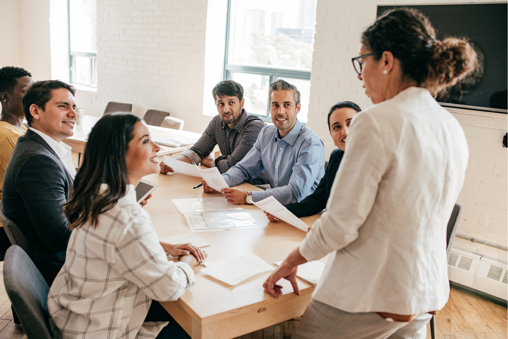 6 persons sitting around a table with documents in front of them and another person standing at one end of the table