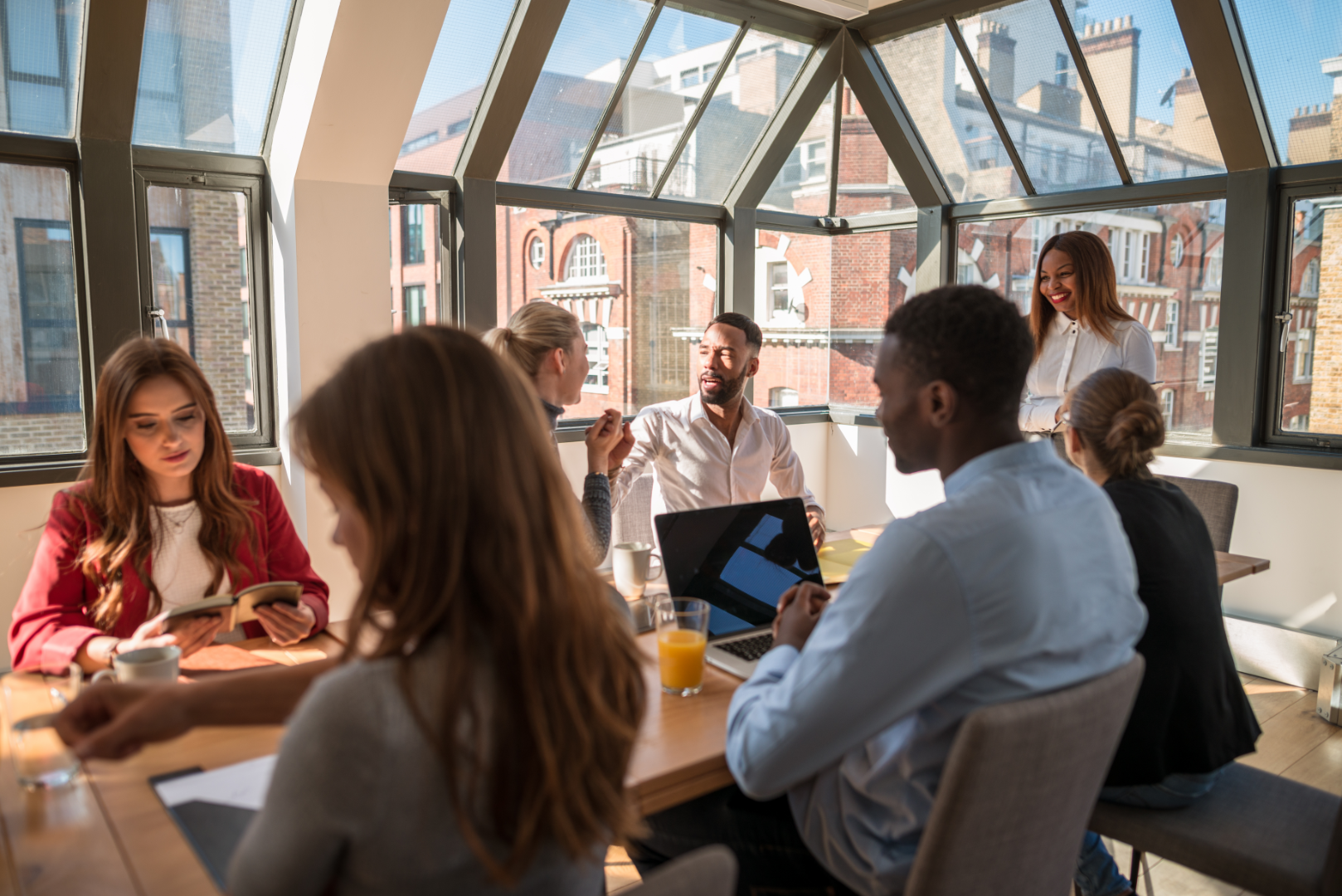 6 persons around a table looking at screens or documents and one person standing at the top of the table. They are in a room with windows showing the buildings around